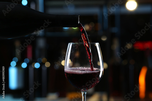 Pouring red wine from bottle into glass on blurred background, closeup photo