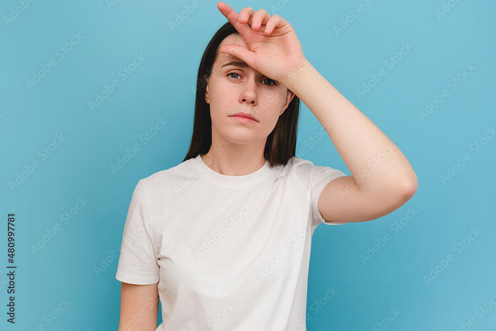 Portrait of tired sad young brunette woman suffering from headache, isolated on blue background with copy space. Cause of headache include migraine, tension headache, stress, depression or brain tumor Portrait of tired sad young brunette woman suffering from headache, isolated on blue background with copy space. Cause of headache include migraine, tension headache, stress, depression or brain tumor
