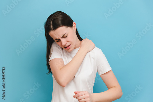 Tired upset young brunette woman massaging hurt stiff neck, fatigued sad brunette girl rubbing tensed muscles to relieve joint shoulder pain, isolated on blue studio background. Fibromyalgia concept photo