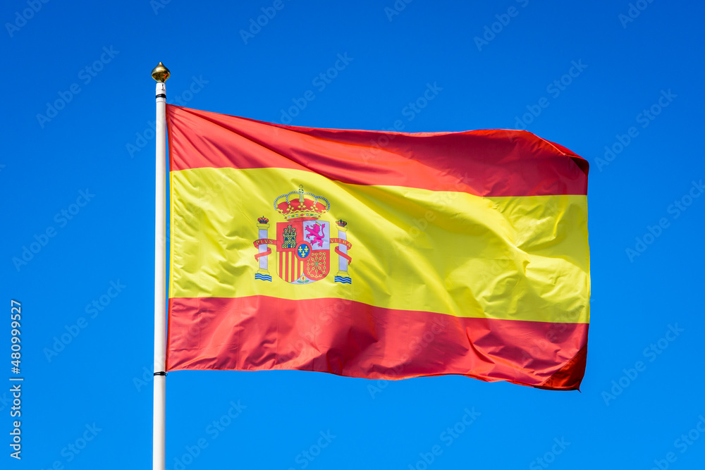 The national flag of Spain is flying in the wind at full mast against blue sky. The national flag of Spain is flying in the wind at full mast against blue sky.