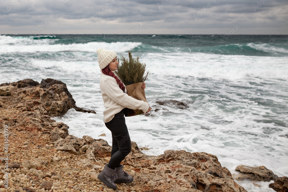 A beautiful girl with long red hair holds a craft bag with Christmas tree branches. A woman is dressed in a white sweater and a hat, she is walking along the winter coast of the sea.