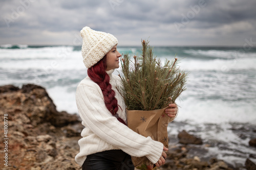 A beautiful girl with long red hair holds a craft bag with Christmas tree branches. A woman is dressed in a white sweater and a hat, she is walking along the winter coast of the sea.