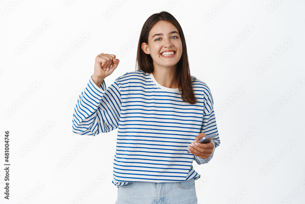 Happy excited young woman holding mobile phone and reacting pleased, winning on smartphone, achieve goal in app, white background Happy excited young woman holding mobile phone and reacting pleased, winning on smartphone, achieve goal in app, white background