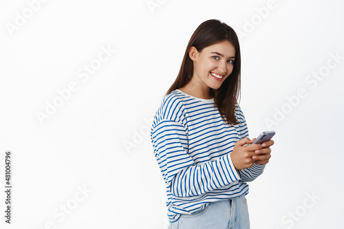 Image of young beautiful woman using mobile phone and looking at camera. Smiling girl holding smartphone on white isolated background