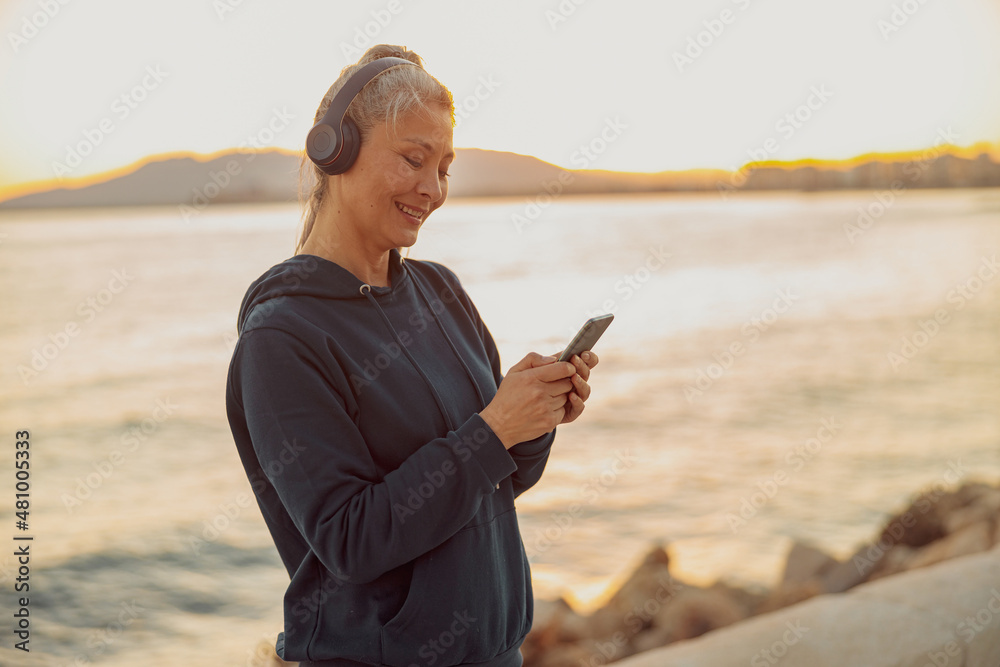 Naklejka premium Charming sportswoman using smartphone and listening to music while standing on background of calm sea and sunset