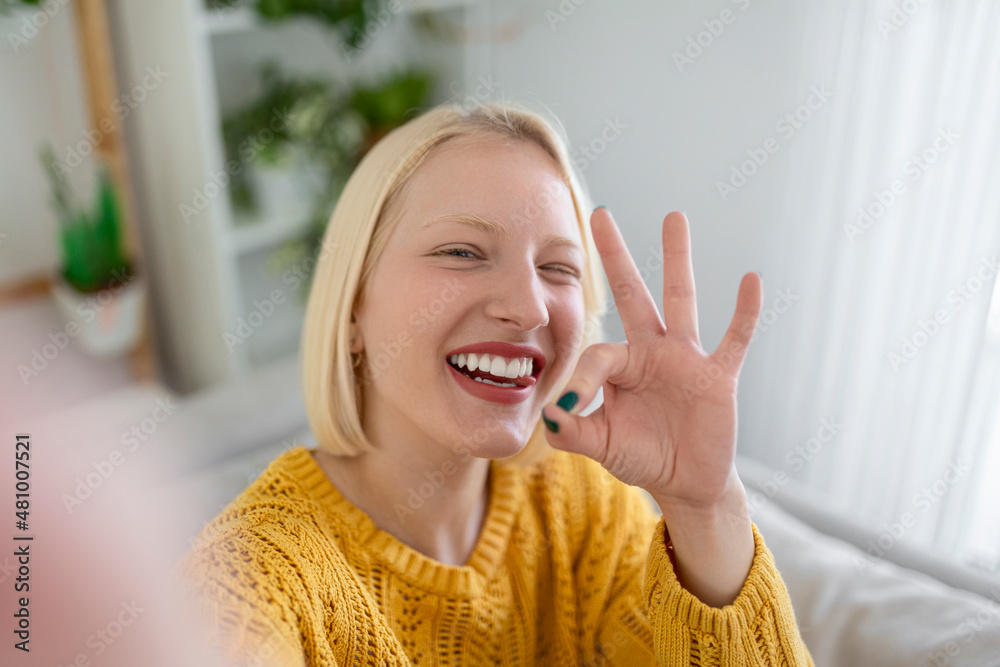 Mischievous young woman takes selfie at home. Beautiful blond woman ok sign with hand blinking with one eye and with her tongue out