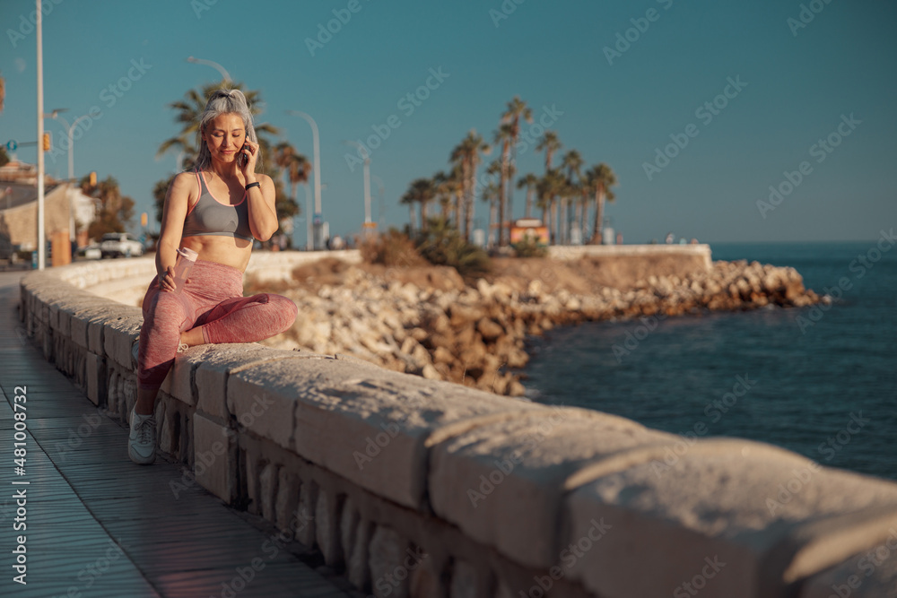 Slender active woman in sports clothes sitting by the seashore, holding mobile phone and talking, looking down