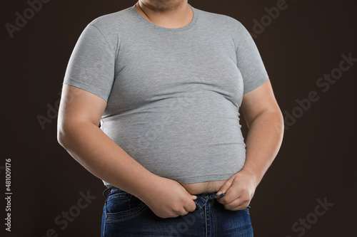 Overweight man trying to button up tight jeans on dark brown background, closeup photo