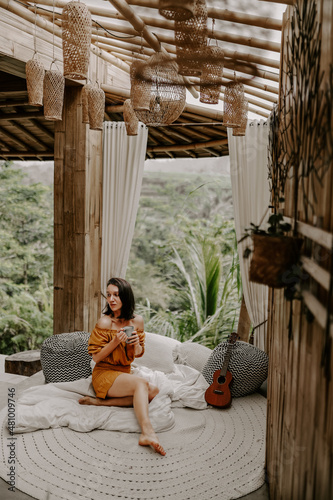 Young female in white shirt is having a relaxing morning drinking coffee in her hotel in Bali, Indonesia, surrounded by green lush landscape