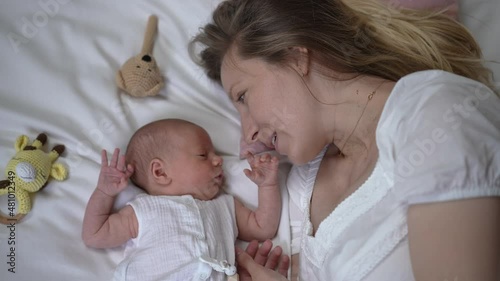 Happy woman talking with newborn boy lying on comfortable soft white bed at home. Top view portrait of excited Caucasian mother admiring infant baby kissing tiny hand in slow motion