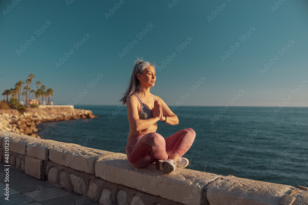 Sporty woman in sports clothes sitting by the beautiful seashore in lotus position and taining on warm day