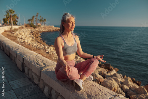 Lady in sports clothes sitting by the beautiful seashore in lotus position and taining on warm day photo