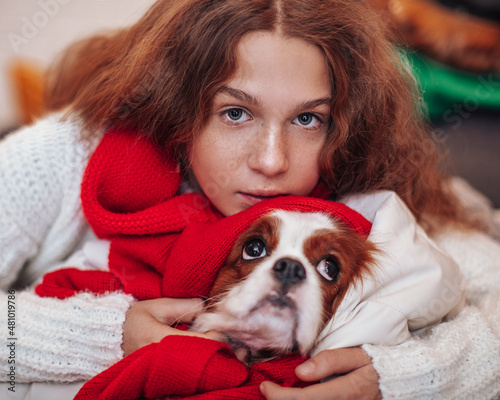 Close up front view of young teen girl with red hair in white sweater and red scarf holding and hugging dog in hands