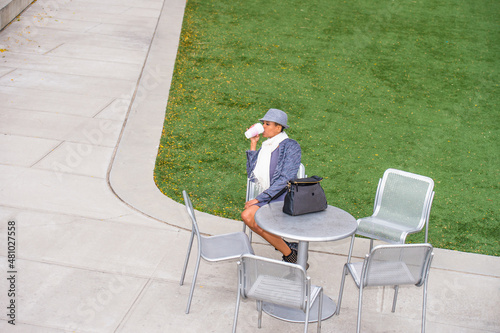 Dressing in faux fur jacket, long white scarf around neck, wearing a woolen Gangster Fedora hat, a young black woman is sitting on a chair by a green lawn, drinking coffee, relaxing, waiting for you