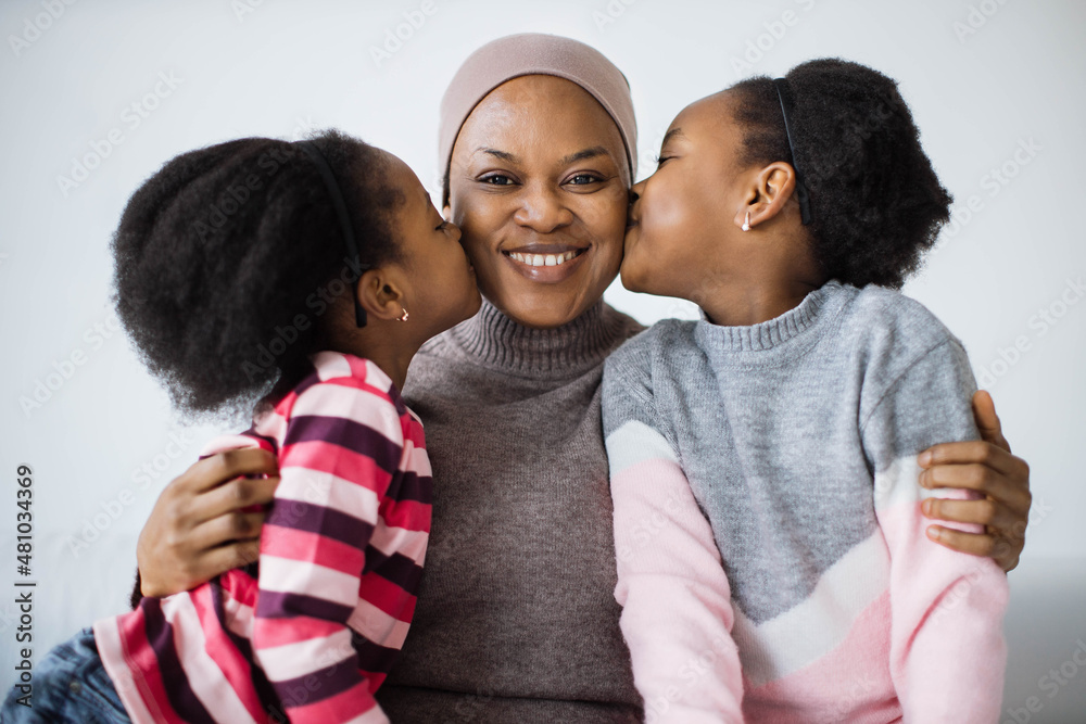 Pretty little sisters kissing mother in cheeks from both sides while sitting together on couch. Happy woman embracing loving daughters and smiling on camera. Pretty little sisters kissing mother in cheeks from both sides while sitting together on couch. Happy woman embracing loving daughters and smiling on camera.