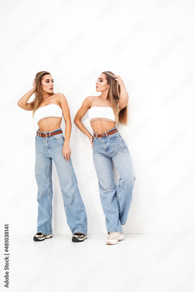 Two beautiful young women with long blond hair and glamour makeup wearing fashionable jeans, standing over white studio background. Sisters. Twins. Two beautiful young women with long blond hair and glamour makeup wearing fashionable jeans, standing over white studio background. Sisters. Twins.