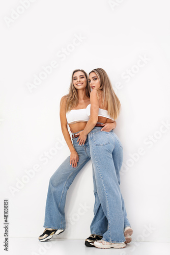Two beautiful young women with long blond hair and glamour makeup wearing fashionable jeans, standing over white studio background. Sisters. Twins.