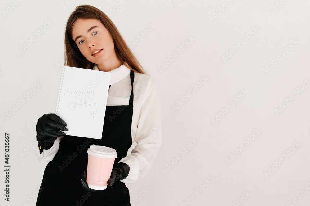 Teenage waitress in a white shirt and black apron holding a notebook, showing it to the camera and a takeaway coffee cup. Morning coffee Teenage waitress in a white shirt and black apron holding a notebook, showing it to the camera and a takeaway coffee cup. Morning coffee
