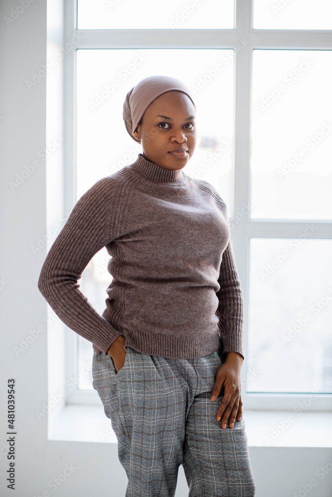 African american muslim woman in headscarf looking at camera while standing near window in white studio. Portrait of middle aged female person. - obrazy, fototapety, plakaty 