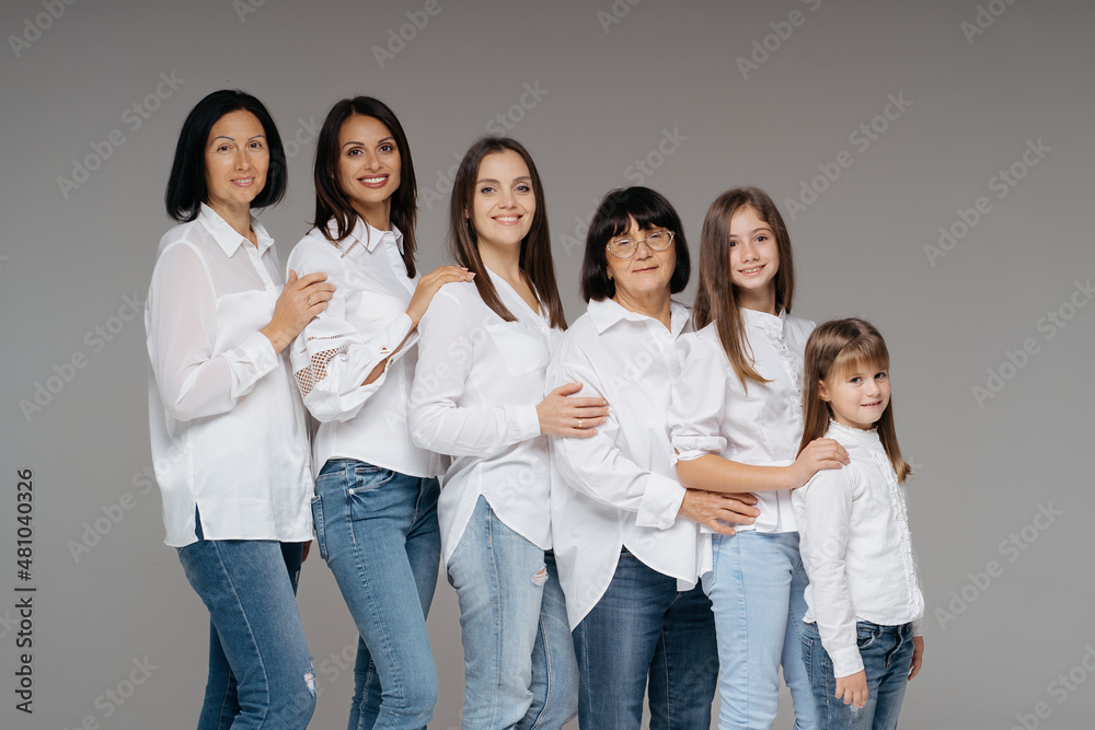 Delighted cute young girls, their moms and grandmother smiling, standing on gray background wearing jeans and white shirts. Lovely women group Delighted cute young girls, their moms and grandmother smiling, standing on gray background wearing jeans and white shirts. Lovely women group