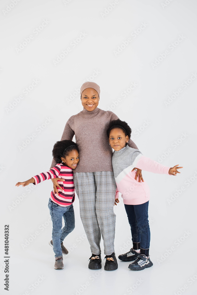 Fototapeta premium Full length portrait of happy african muslim woman in hijab embracing her smiling daughters while standing over white background. Family leisure and playful mood concept.