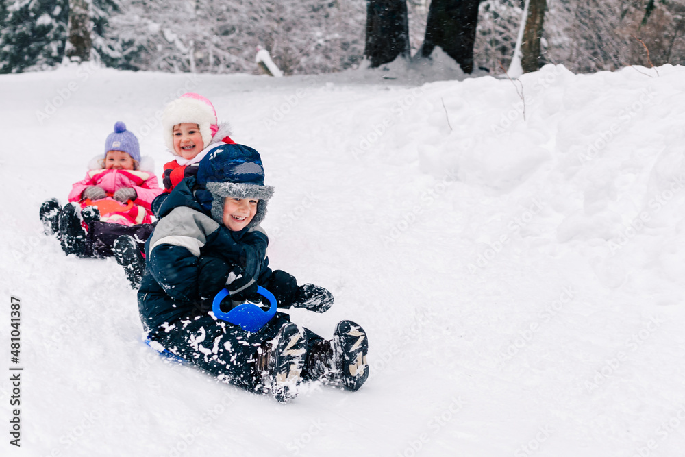 Little caucasian kids in bright clothing laughing and toboganning on snow covered hill. Full lengh horizontal shot. Selective focus on one boy. Happy childhood and active wintertime concept. Little caucasian kids in bright clothing laughing and toboganning on snow covered hill. Full lengh horizontal shot. Selective focus on one boy. Happy childhood and active wintertime concept.