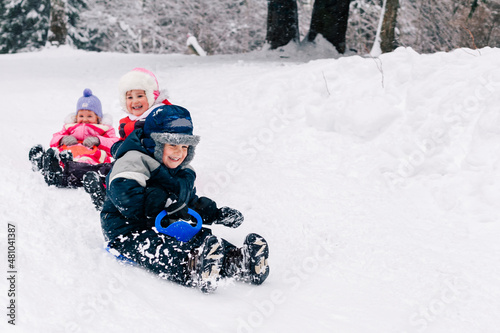 Little caucasian kids in bright clothing laughing and toboganning on snow covered hill. Full lengh horizontal shot. Selective focus on one boy. Happy childhood and active wintertime concept.