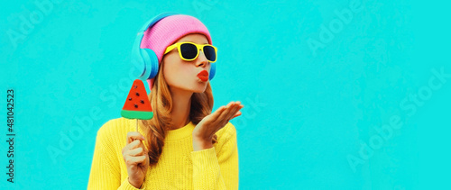 Summer fresh colorful portrait of happy young woman in headphones listening to music with fruit juicy lollipop or ice cream shaped slice of watermelon on blue background