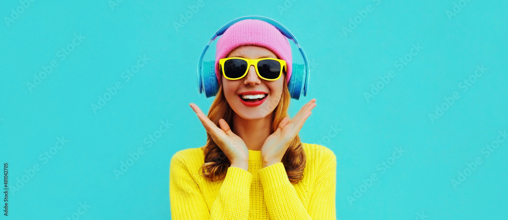 Portrait of happy surprised smiling young woman model with headphones listening to music wearing colorful yellow sweater on blue background