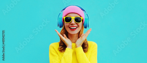 Portrait of happy surprised smiling young woman model with headphones listening to music wearing colorful yellow sweater on blue background