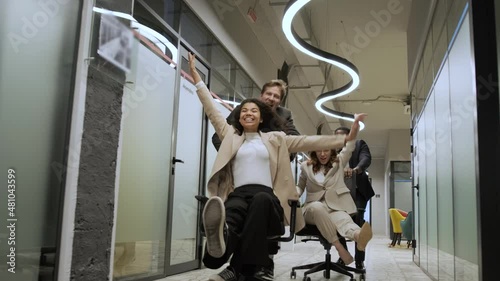 Millennial multiracial team people having fun riding on chairs in office room, excited diverse employees laughing enjoying funny activity at work break, creative friendly workers play game together
