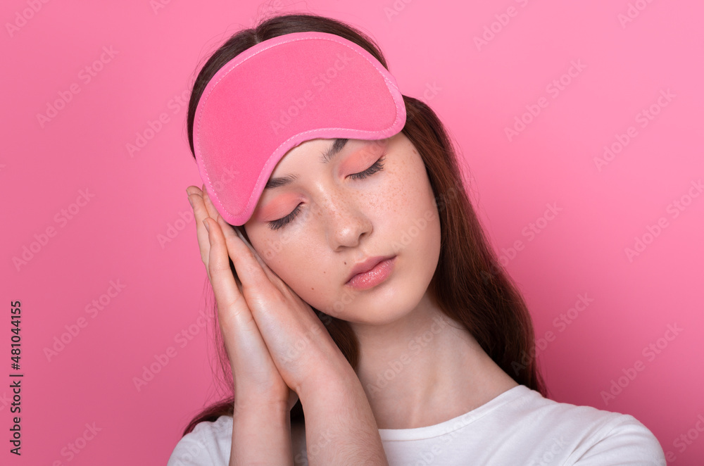 lovely sleepy calm caucasian brunette teen girl with freckles and closed eyes folded palms under head wearing sleep mask and white t-shirt isolated on pink background close-up portrait