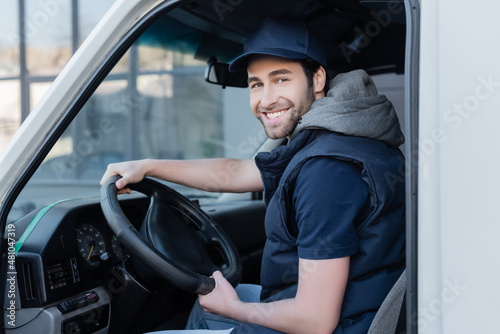 Smiling delivery man looking at camera near steering wheel in car.