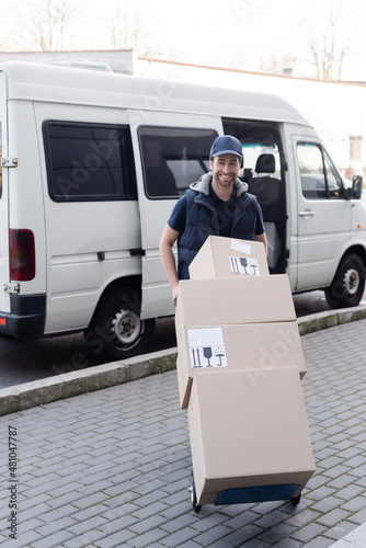 Smiling courier in cap holding cart with boxes near car outdoors. © LIGHTFIELD STUDIOS