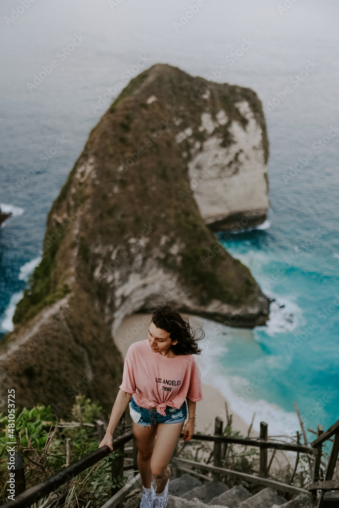 A young travelling and backpacking girl visits Kelingking Beach in Nusa Penida, Indonesia. Overlooking crystal clear turquoise water, waves, blue ocean and sky on a sunny day.