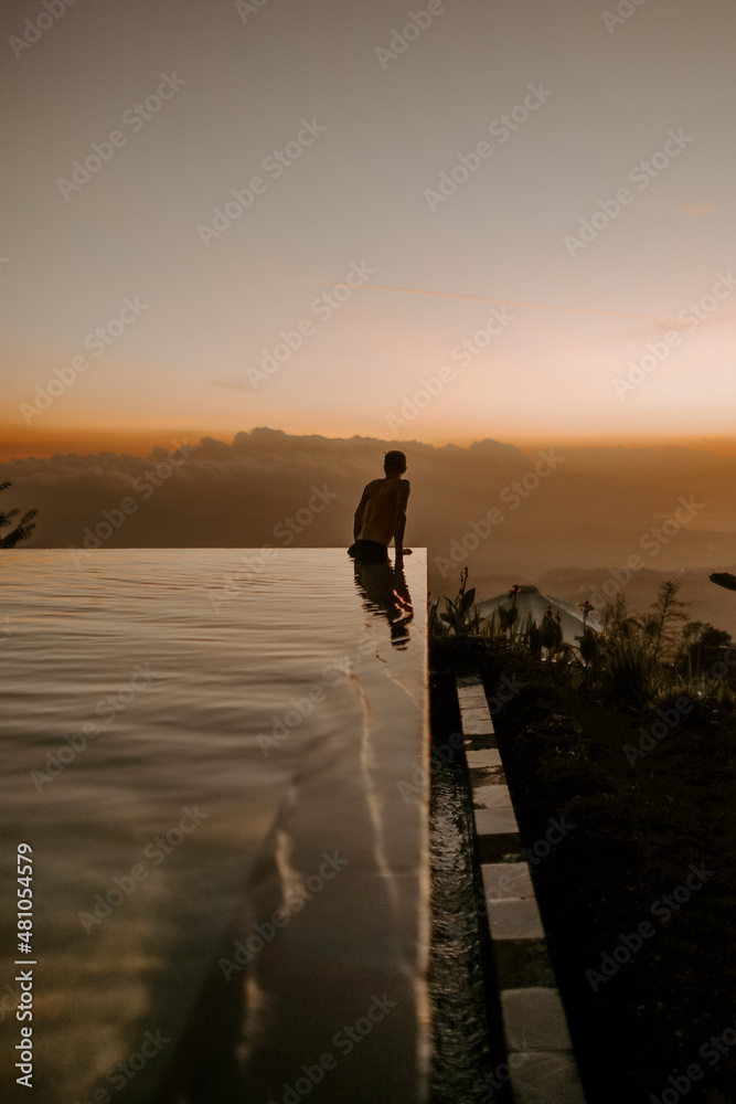 A young male traveller and blogger is walking on the edge of infinity pool at a luxury jungle resort in Bali, Indonesia at sunset.