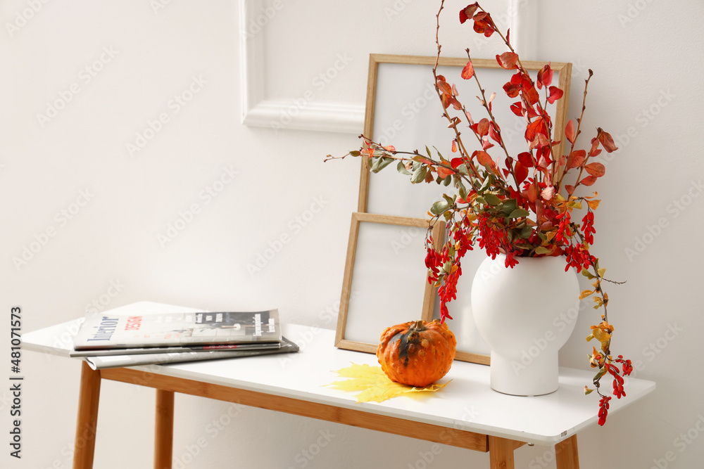 Stylish vase with branches, blank frames, magazines and pumpkin on table near white wall - obrazy, fototapety, plakaty 