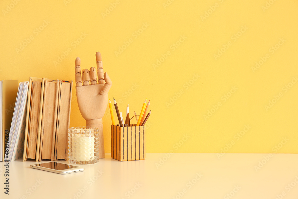 Books, wooden mannequin hand, stationery holder and mobile phone on table near color wall - obrazy, fototapety, plakaty 