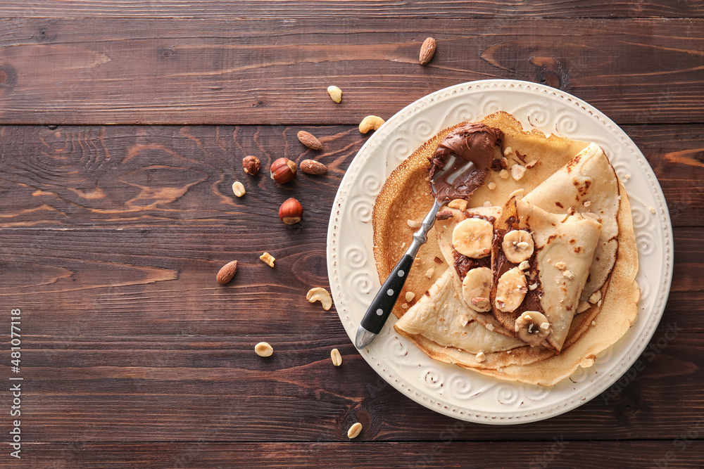 Plate of tasty thin pancakes with chocolate paste, cut banana and nuts on wooden background