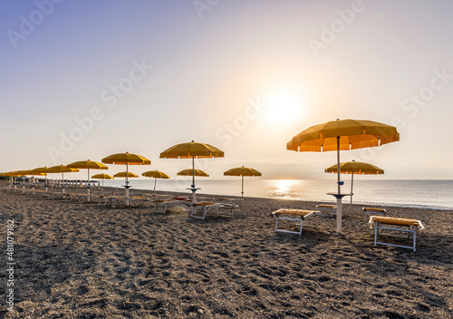 scenic view at nice beach with rows of umbrellas and chaise lounges with blue sea and amazing sunrise sky with sun glow on the background