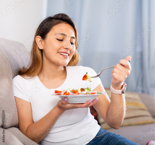 Positive peruvian housewoman eating vegetable salad on sofa at home.. photo