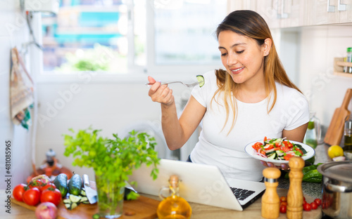 Young peruvian housewoman eating vegetable salad and watching movie on laptop photo