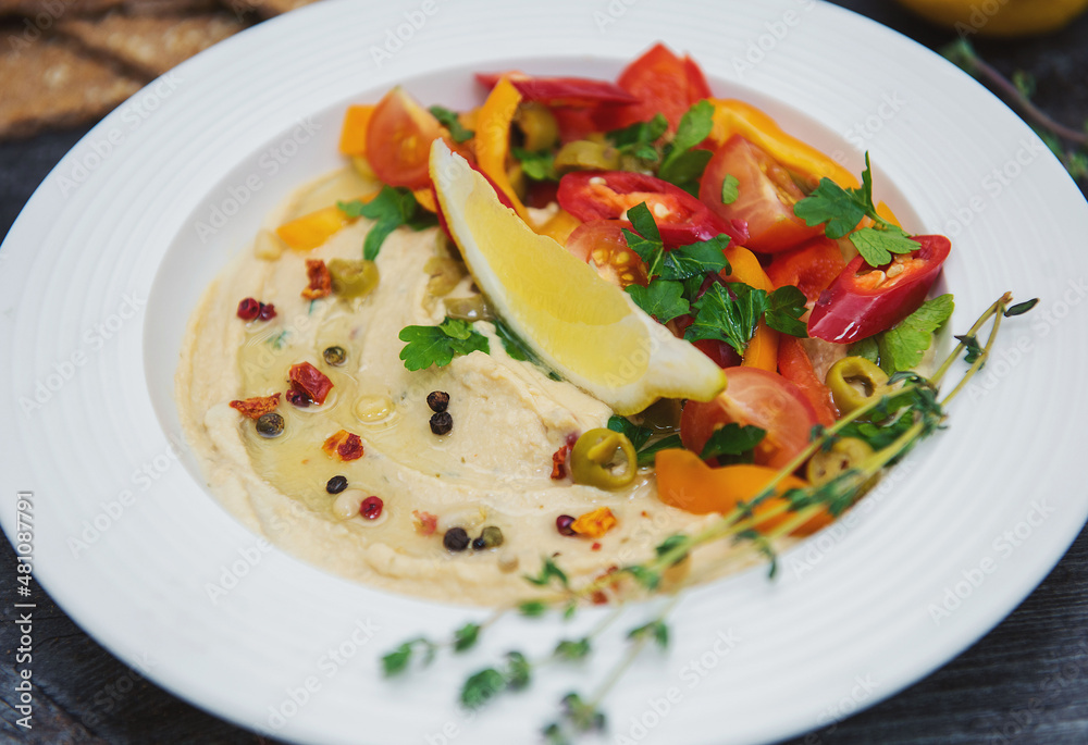 in a white plate hummus with a slice of lemon chopped hot pepper slices of tomato and spicy seasoning with sprigs of greenery on the edge in a white plate hummus with a slice of lemon chopped hot pepper slices of tomato and spicy seasoning with sprigs of greenery on the edge