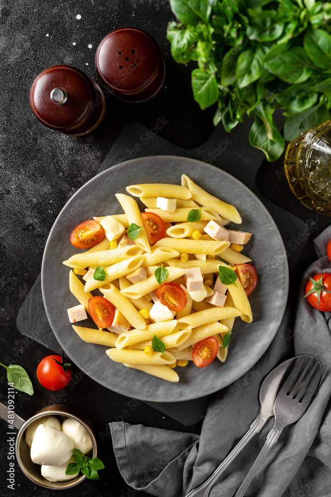 Pasta salad with ham, mozzarella, corn, tomatoes and olive oil in a ceramic plate on a dark culinary background. Traditional Italian dish penne rigate with cheese and vegetables top view