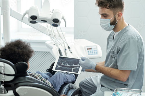 Dentist in mask, gloves and uniform interacting with little patient and showing him x-ray of teeth on tablet display