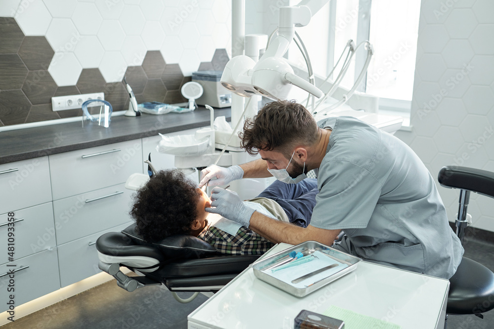 Contemporary dentist bending over little patient with cavity while