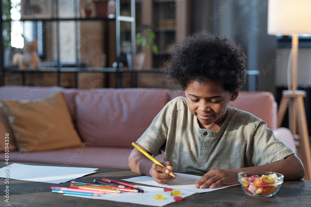 Naklejka premium Happy schoolboy of African ethnicity drawing picture with crayons while sitting by table in home environment