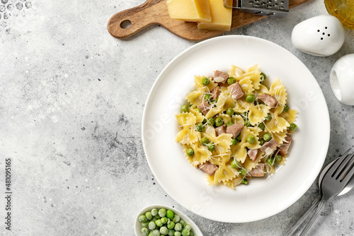 Farfalle with green peas, turkey ham and parmesan in cream sauce in a white plate on the kitchen table. Traditional Italian pasta on a light culinary background. Top view	