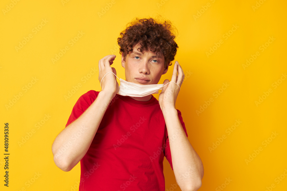 Young curly-haired man wearing a red t-shirt medical mask on the face posing isolated background unaltered Young curly-haired man wearing a red t-shirt medical mask on the face posing isolated background unaltered