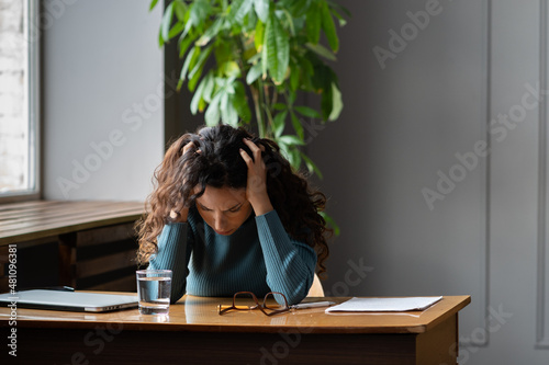 Anxiety at work. Young stressed businesswoman holding head in hands having headache or migraine, sitting at office desk with water glass, tired female employee suffering from hypertension at workplace
